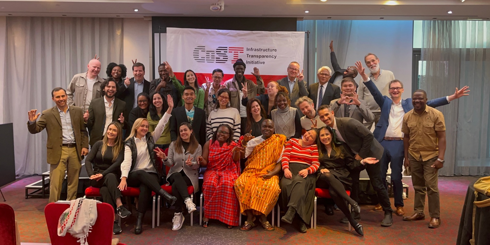 CoST members, partners and Secretariat staff together at a CoST gathering, waving in front of the CoST banner